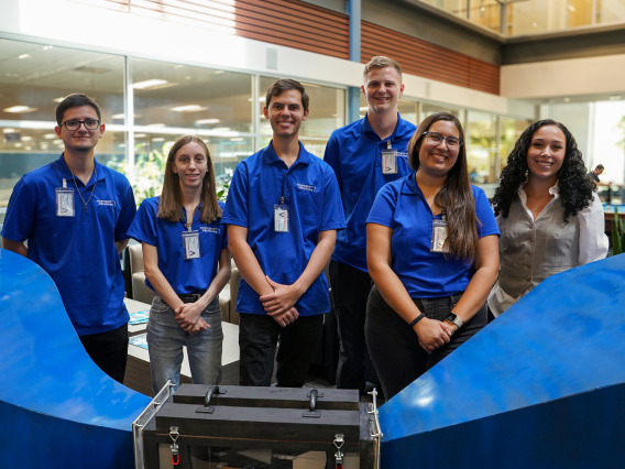 Five people in blue shirts pose for a photo with a woman in a tan vest.