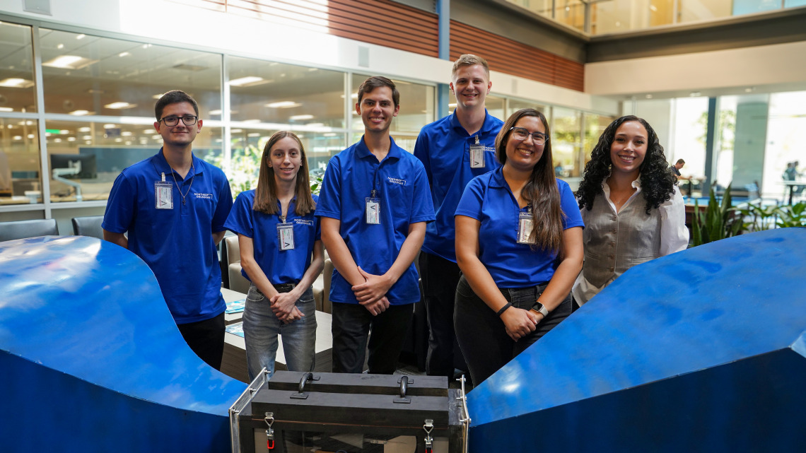 Five people in blue shirts pose for a photo with a woman in a tan vest.