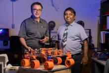 Jekan Thanga (right) with collaborator Moe Momayez in a lab. They are standing behind a small orange robot with six wheels.