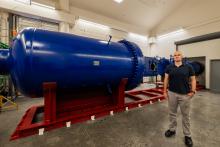 Jesse Little stands next to a wind tunnel, a large blue horizontal cylinder.