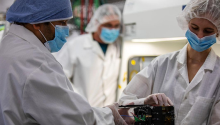 Three University of Arizona students work on a satellite while wearing lab coats and face masks