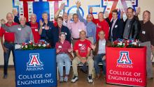 About 15 members of the Class of 1969 stand together in the foyer of the Student Union Memorial Center Ballroom. Most are wearing red or blue shirts. Their hands are raised to wave at the camera.