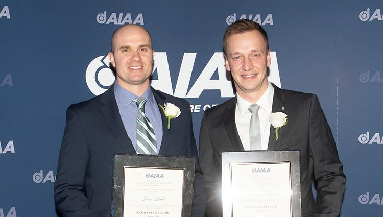 Two men in dark suits stand in front of an AIAA-branded background. They are holding their associate fellow certificates.