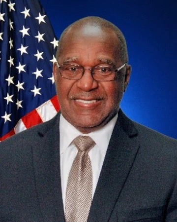 A man with dark skin wears circle glasses and poses for a photo.