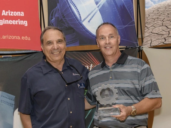 Two older men pose for a photo. The man on the left wears a dark navy shirt and the man on the right wears a grey striped shirt.