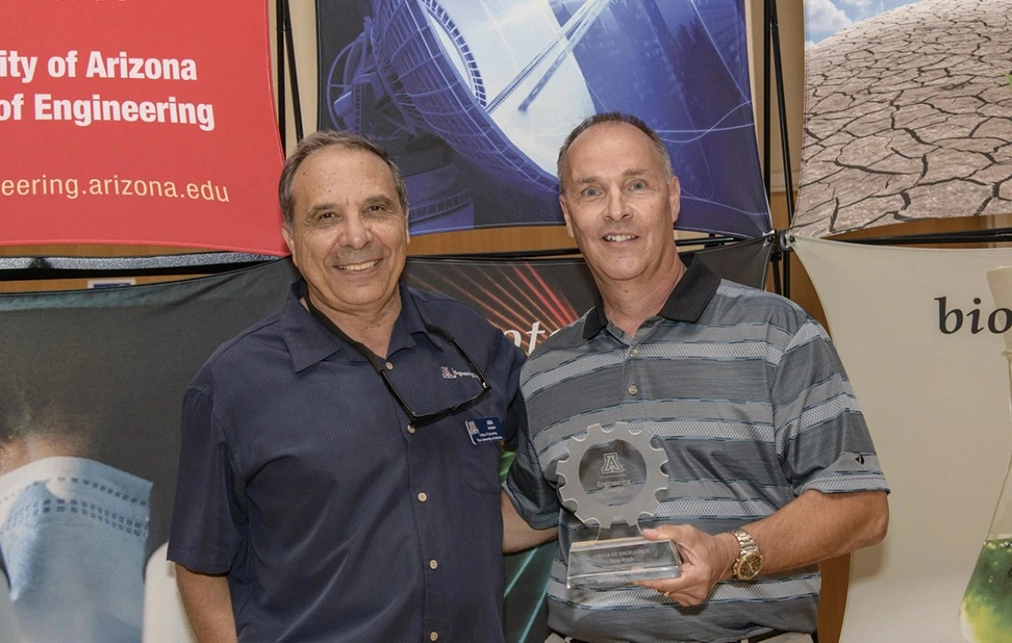 Two older men pose for a photo. The man on the left wears a dark navy shirt and the man on the right wears a grey striped shirt.